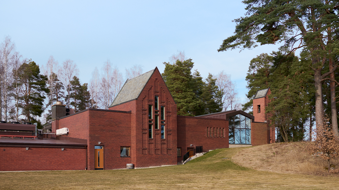En röd tegelbyggnad med höga fönster och brant tak står på en gräskulle, omgiven av träd och ett glest skogsparti.