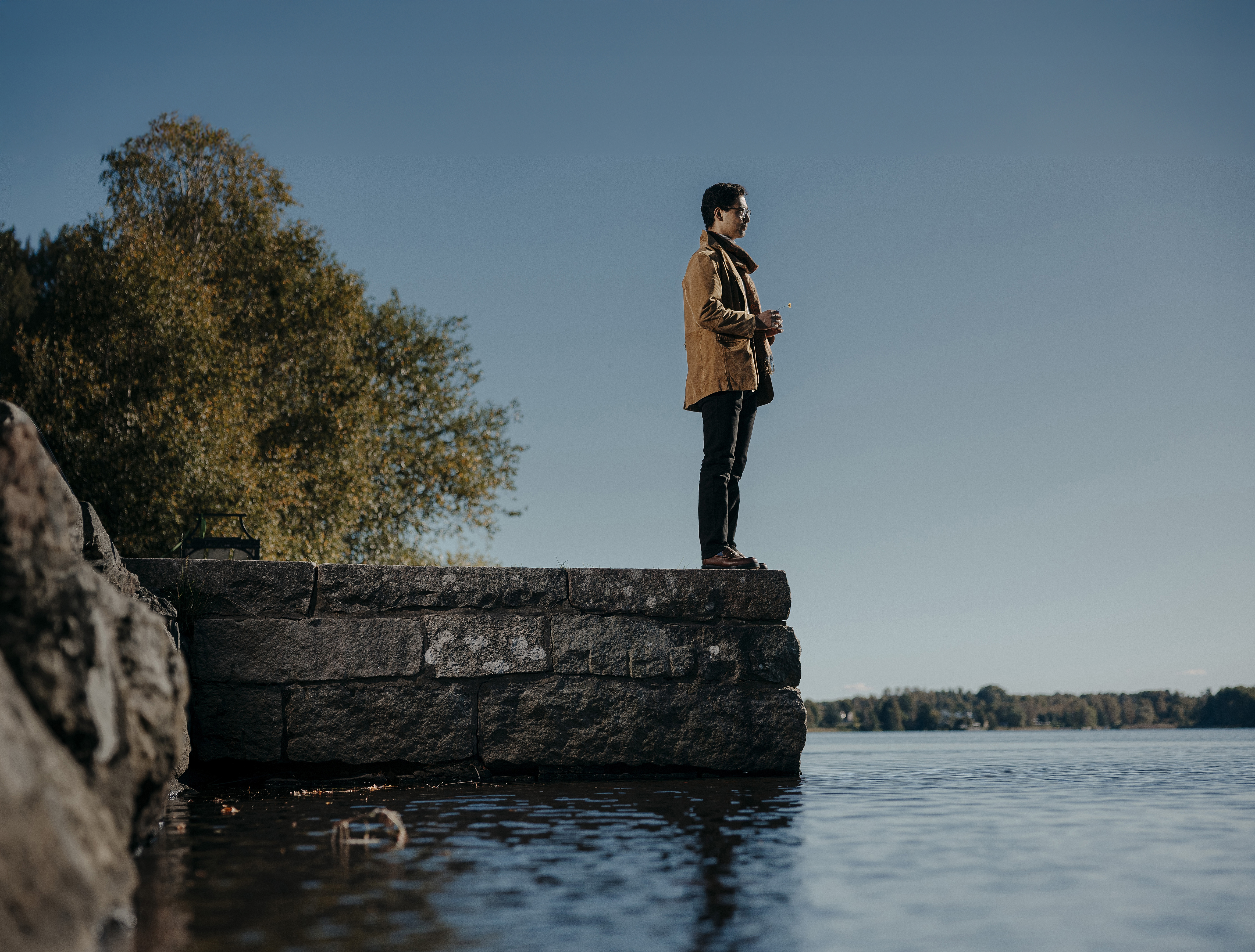 En person står vid kanten av en stenpir och ser ut över en sjö, med träd och blå himmel i bakgrunden.