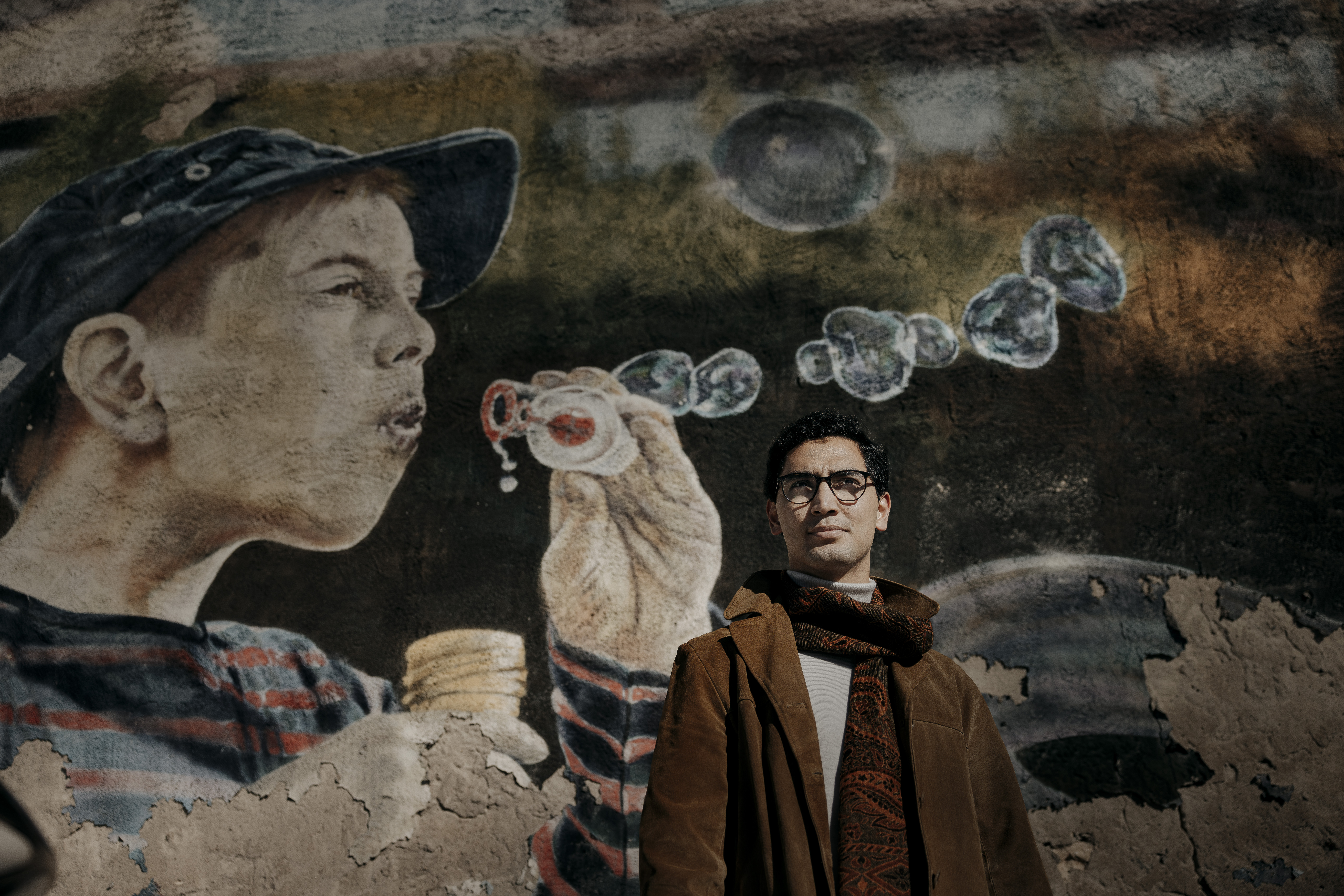 En man med glasögon och brun rock står framför en väggmålning som visar en pojke som blåser såpbubblor.