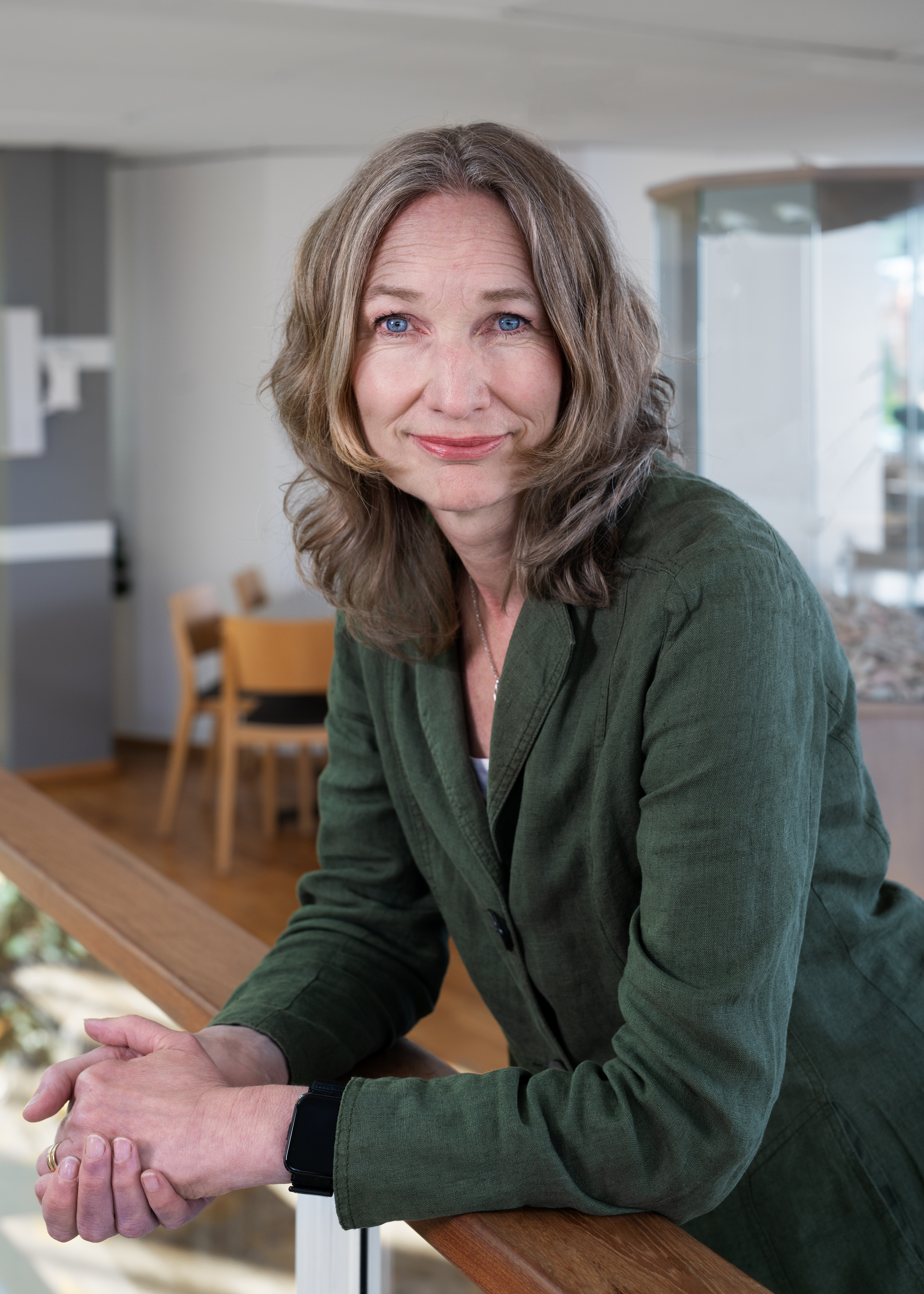 En kvinna med vågigt, axellångt hår och grön kavaj lutar sig mot ett träräcke inomhus och tittar in i kameran.