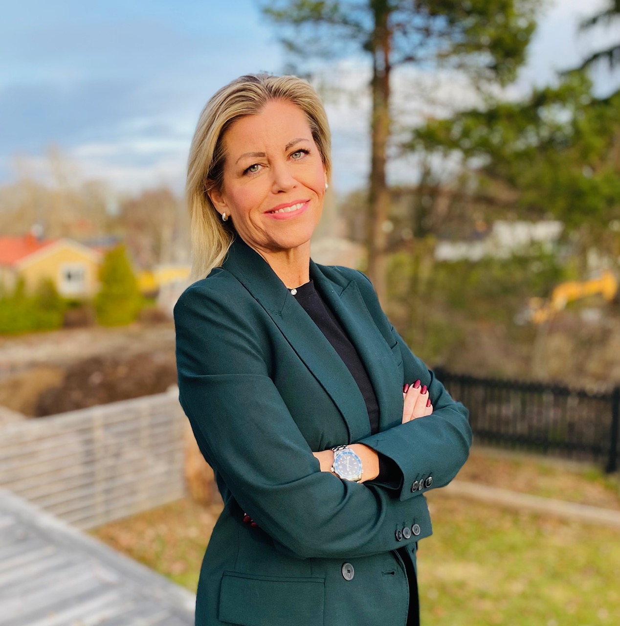 En kvinna i mörkgrön kavaj står utomhus med korsade armar och ler mot kameran. Träd och byggnader syns i bakgrunden.