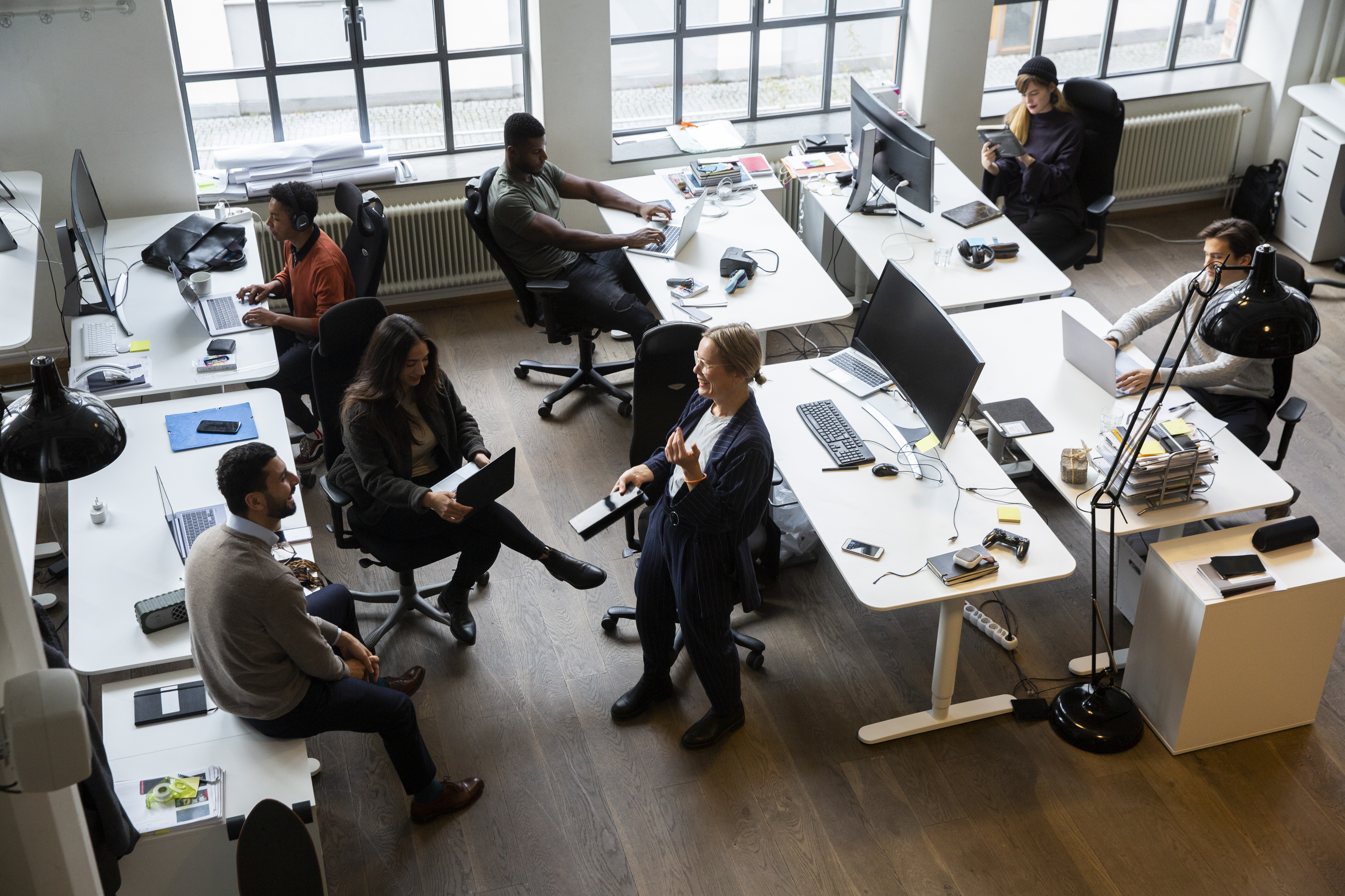 Sex personer arbetar i ett öppet kontorslandskap; några sitter vid datorer, andra står och pratar. Ljus kommer in genom fönster.