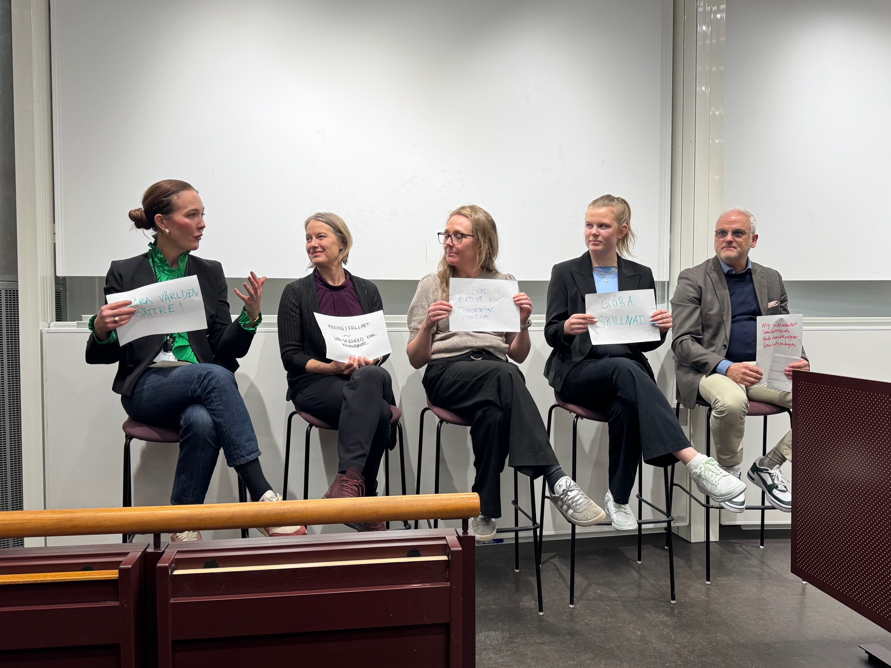 Fem personer sitter på pallar framför en whiteboard, håller varsitt papper med text och deltar i en paneldiskussion.