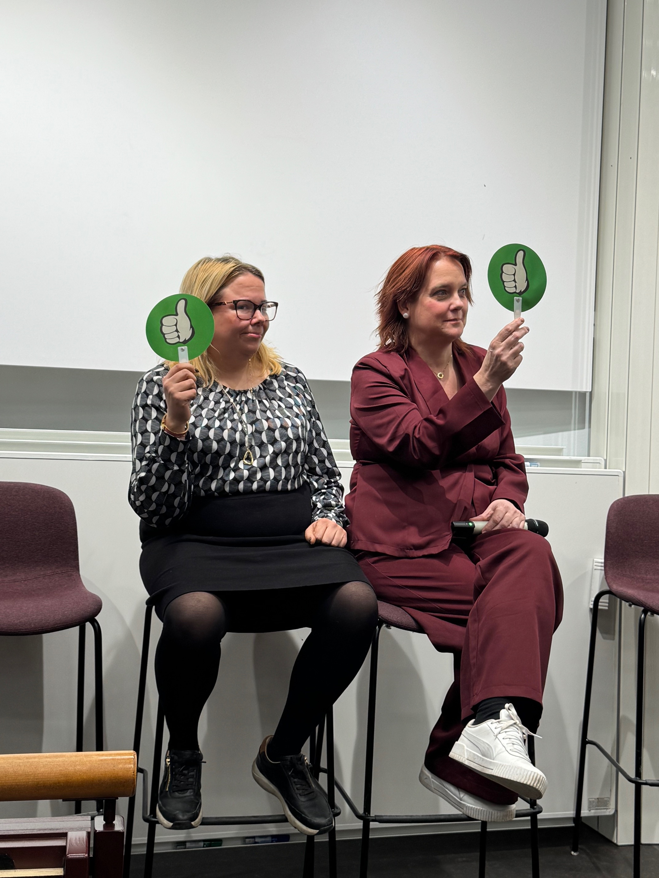 Två kvinnor sitter på pallar och håller upp gröna skyltar med en tumme upp, under en paneldiskussion eller gruppdiskussion.