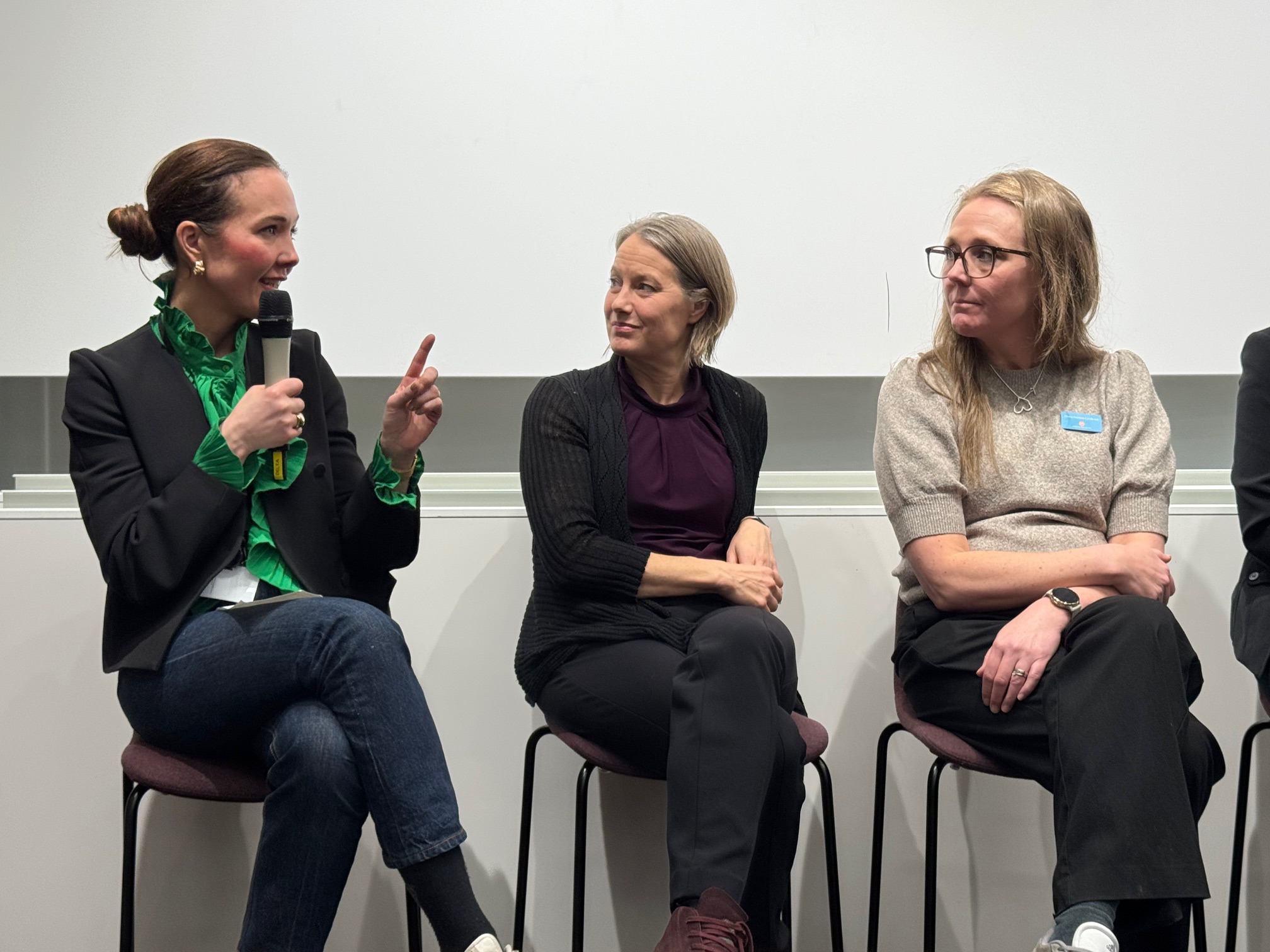 Tre kvinnor sitter på stolar under en paneldiskussion; kvinnan till vänster talar i en mikrofon, de andra två lyssnar.