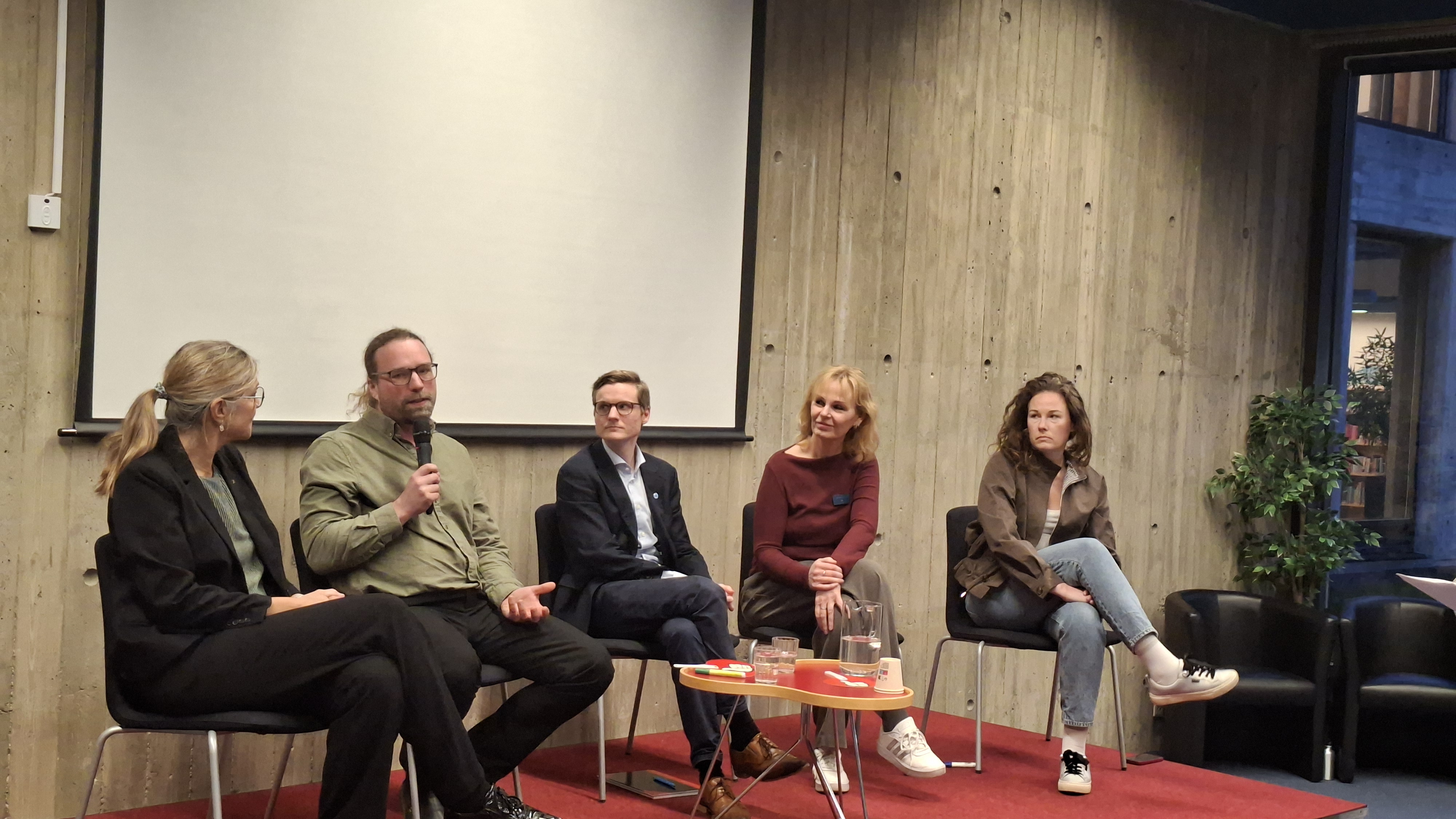 Fem personer sitter på stolar framför en tom projektorduk, en person talar i mikrofon under en paneldiskussion i ett rum.