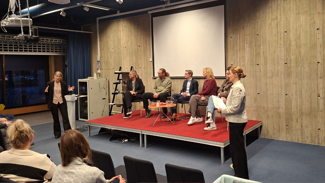 Fem personer sitter på en liten scen och en person står och talar till en publik i ett konferensrum.
