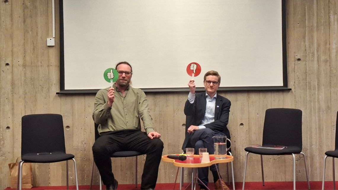 Två män sitter på en scen och håller upp paddlar; mannen till vänster visar en grön tumme upp, mannen till höger en röd tumme ner. En tom stol och ett bord står mellan dem.