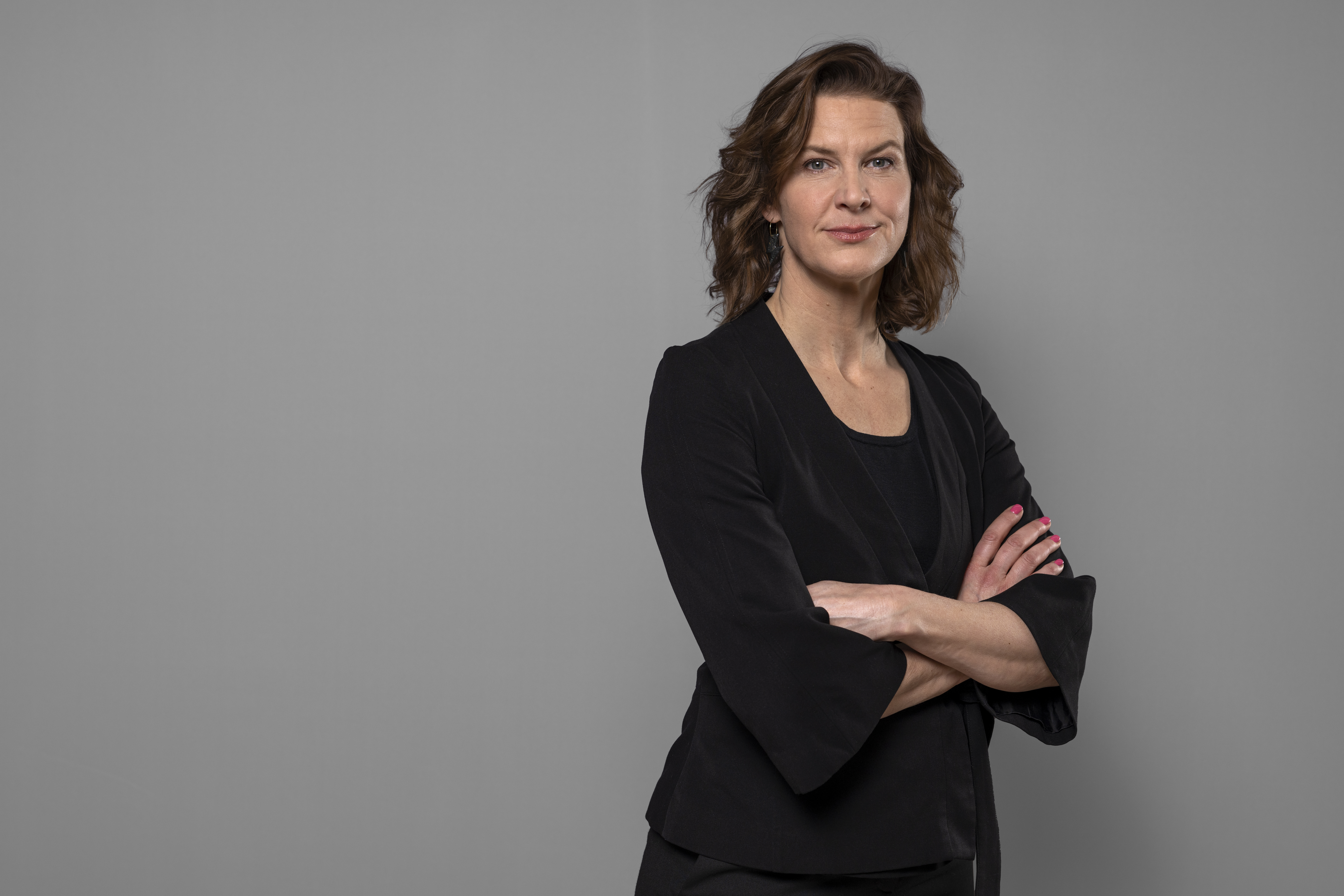 Kvinna med brunt hår har svart kavaj och står med armarna i kors mot en grå bakgrund.