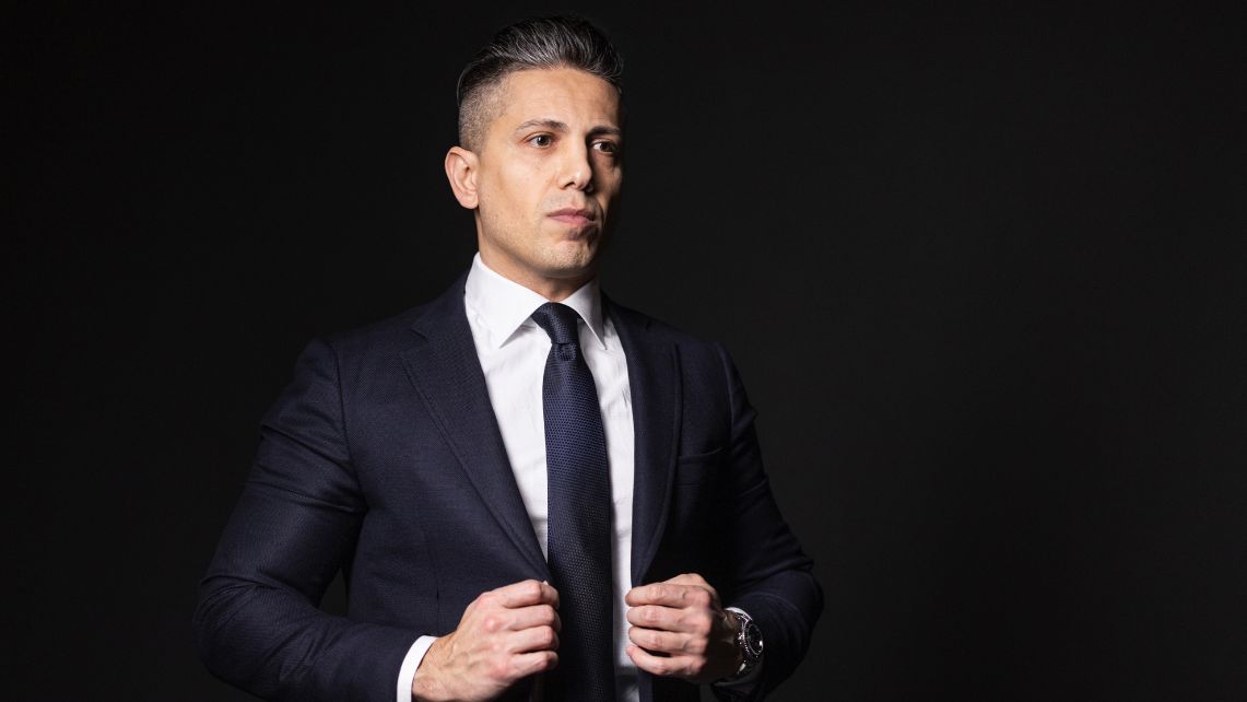 En man i kostym och slips står mot en svart bakgrund, justerar sin kavaj med båda händerna och tittar framåt.