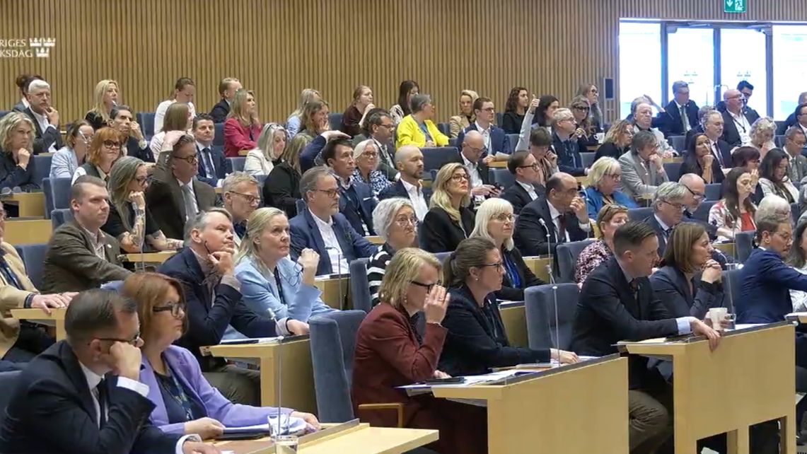 En grupp personer sitter i en träpanelad hörsal, vända framåt, med papper och enheter på borden.