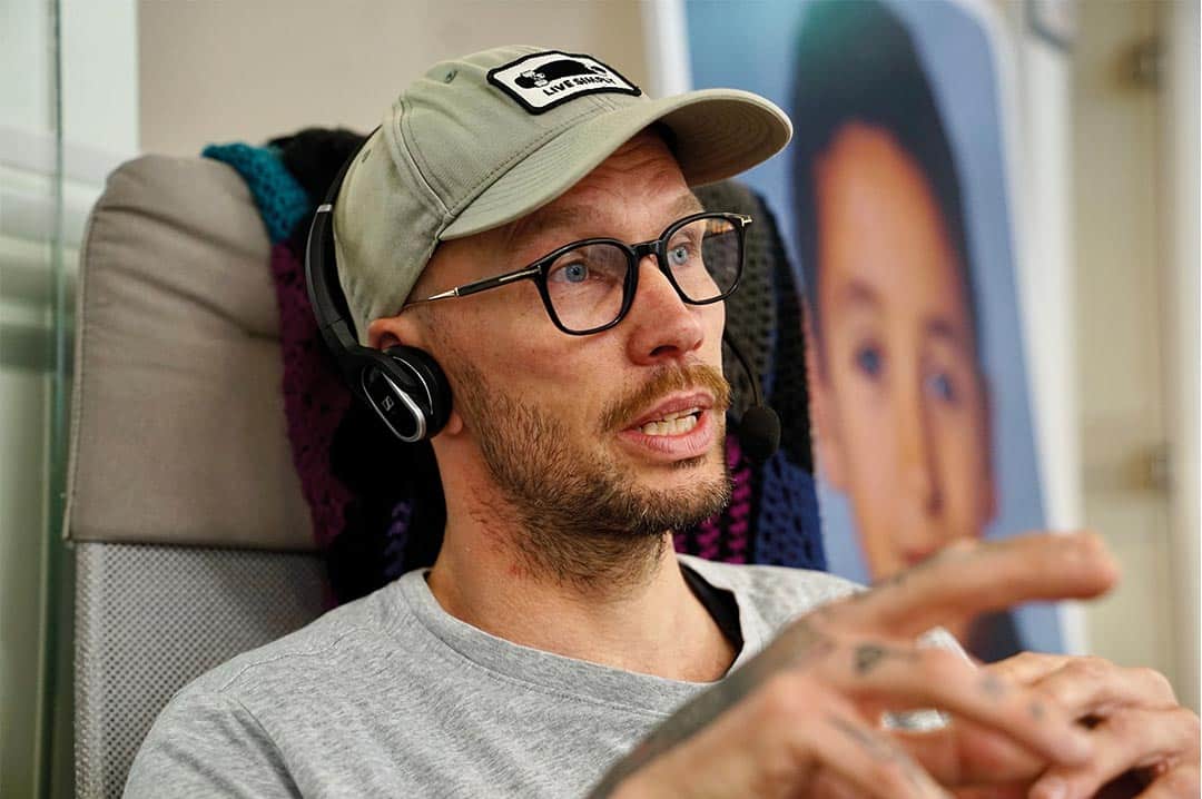 Jonas Söderlund med glasögon, keps och headset sitter inomhus och talar medan han gestikulerar.