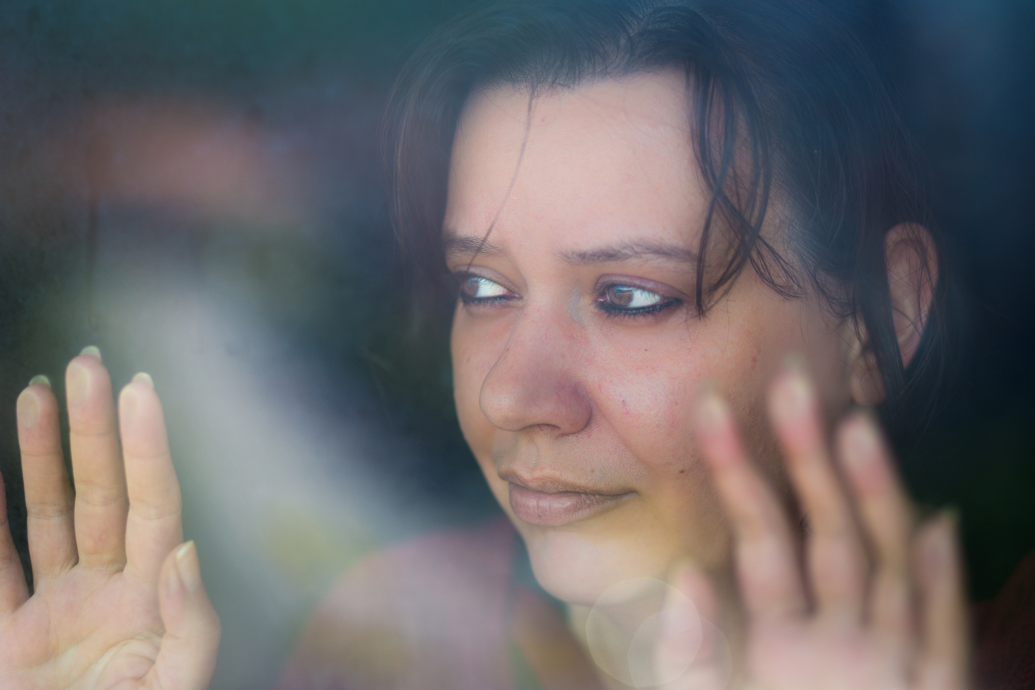 Close up color image depicting a sad, depressed-looking woman in her 30s, and of caucasian ethnicity, looking out the window with her hands pressed to the glass. The image was taken during the Covid-19 pandemic during lockdown, and the image illustrates a mental health concept, as well as a domestic abuse concept. Room for copy space.