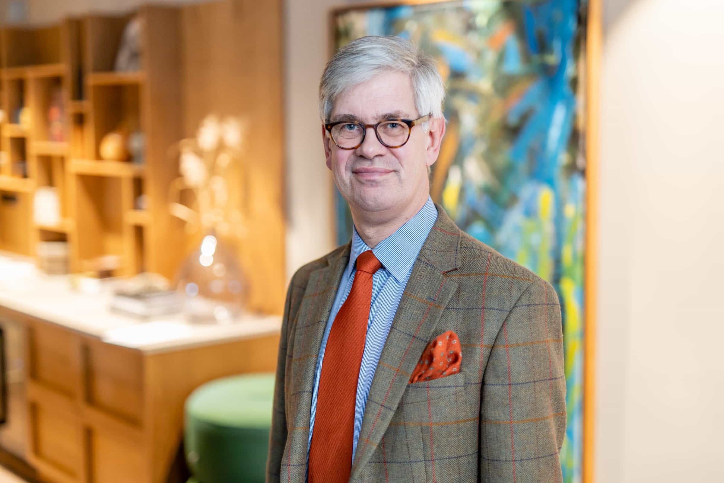 En man med grått hår och glasögon, klädd i rutig kavaj, blå skjorta och orange slipsar, står inomhus med hyllor och konst bakom sig.