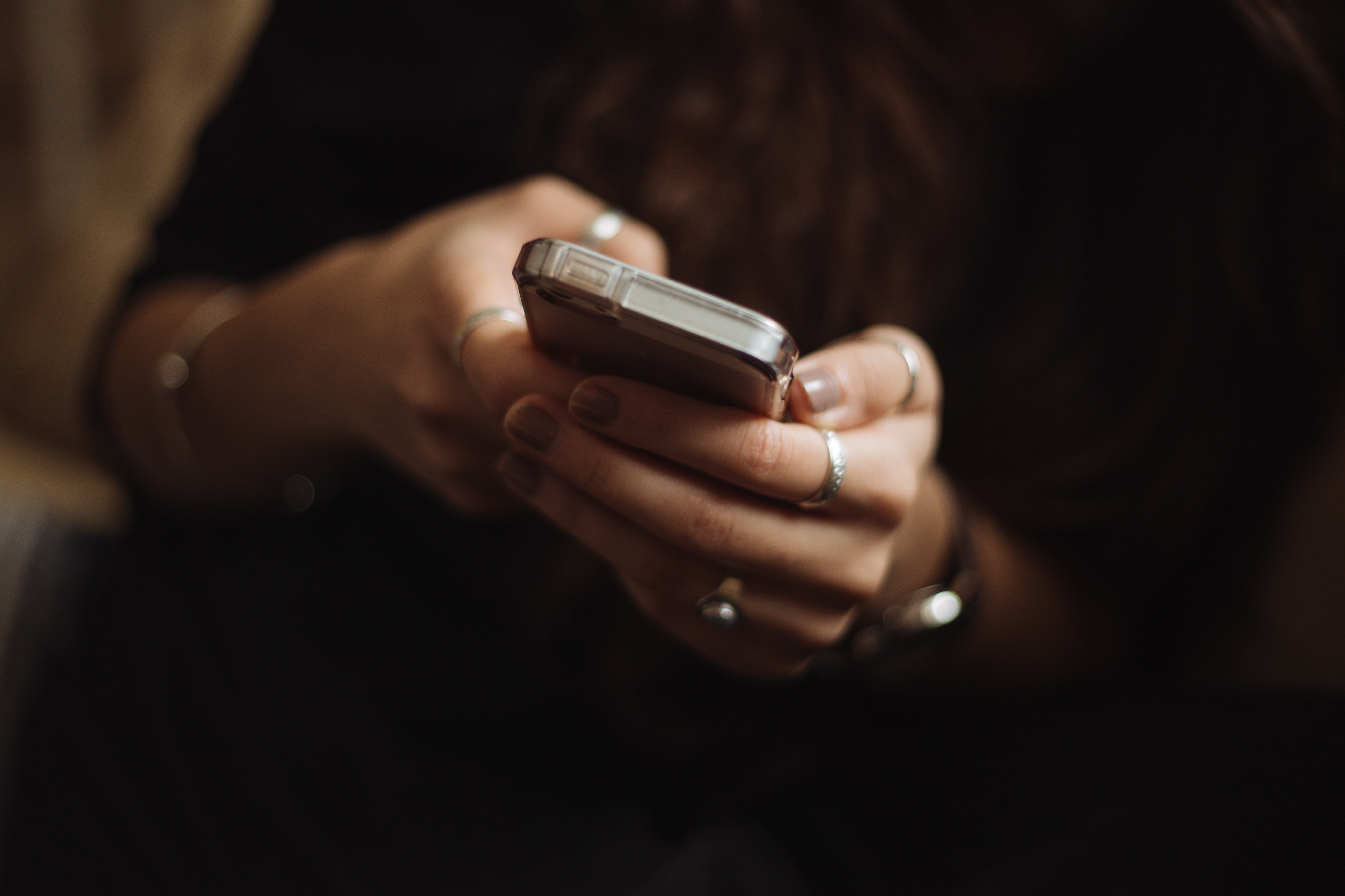 En person med långt hår, med ringar på fingrarna och mörk tröja, håller och använder en smartphone med båda händerna.