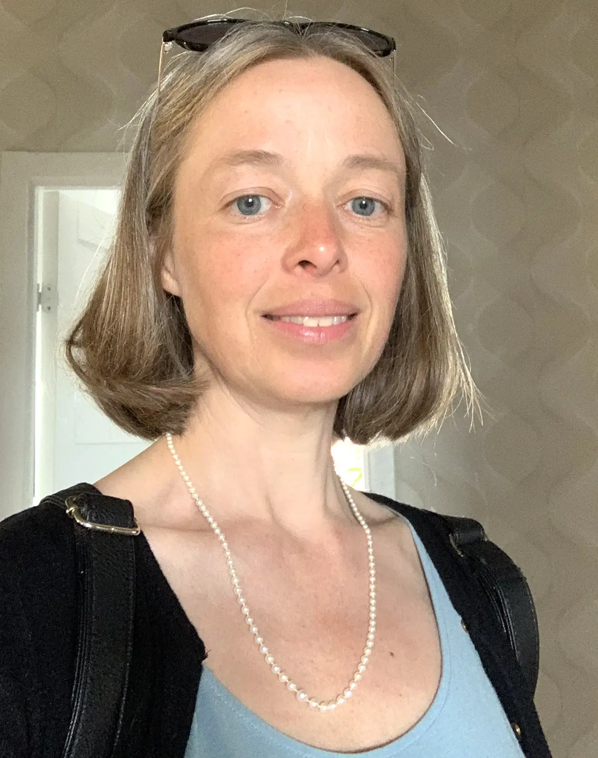 Anna Lundqvist med axellångt ljusbrunt hår, ljusblå topp, svart kofta, pärlhalsband och solglasögon står inomhus och ler mot kameran.