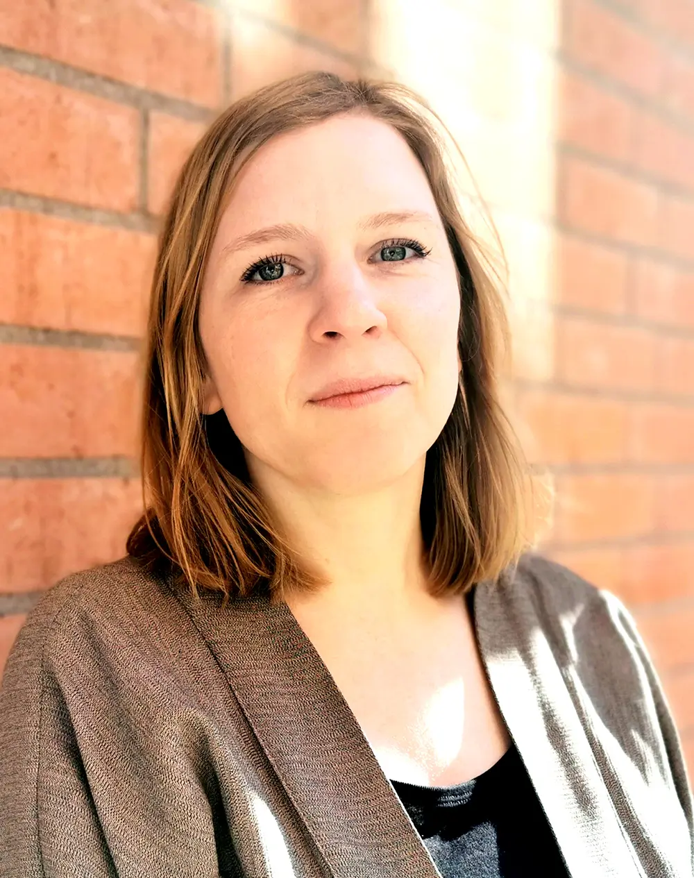 Kerstin Annerborn med axellångt ljust brunt hår står mot en tegelvägg, har en brun jacka över en svart topp och ser in i kameran.