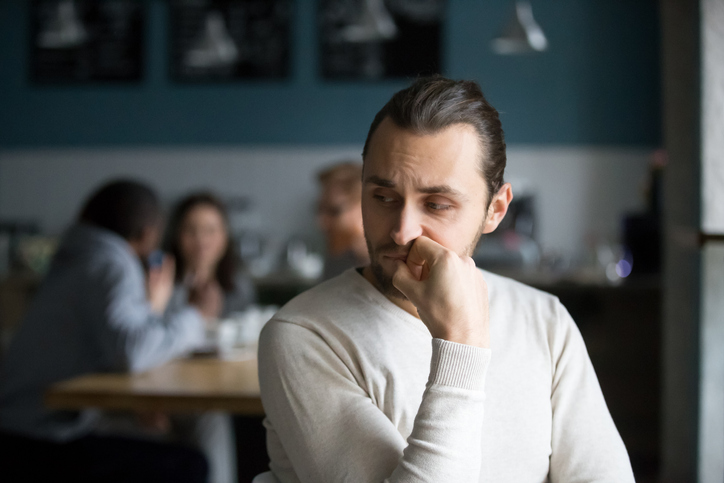 En man i tröja sitter vid ett bord, vilar hakan i handen och tittar åt sidan, med en grupp personer suddiga i bakgrunden.