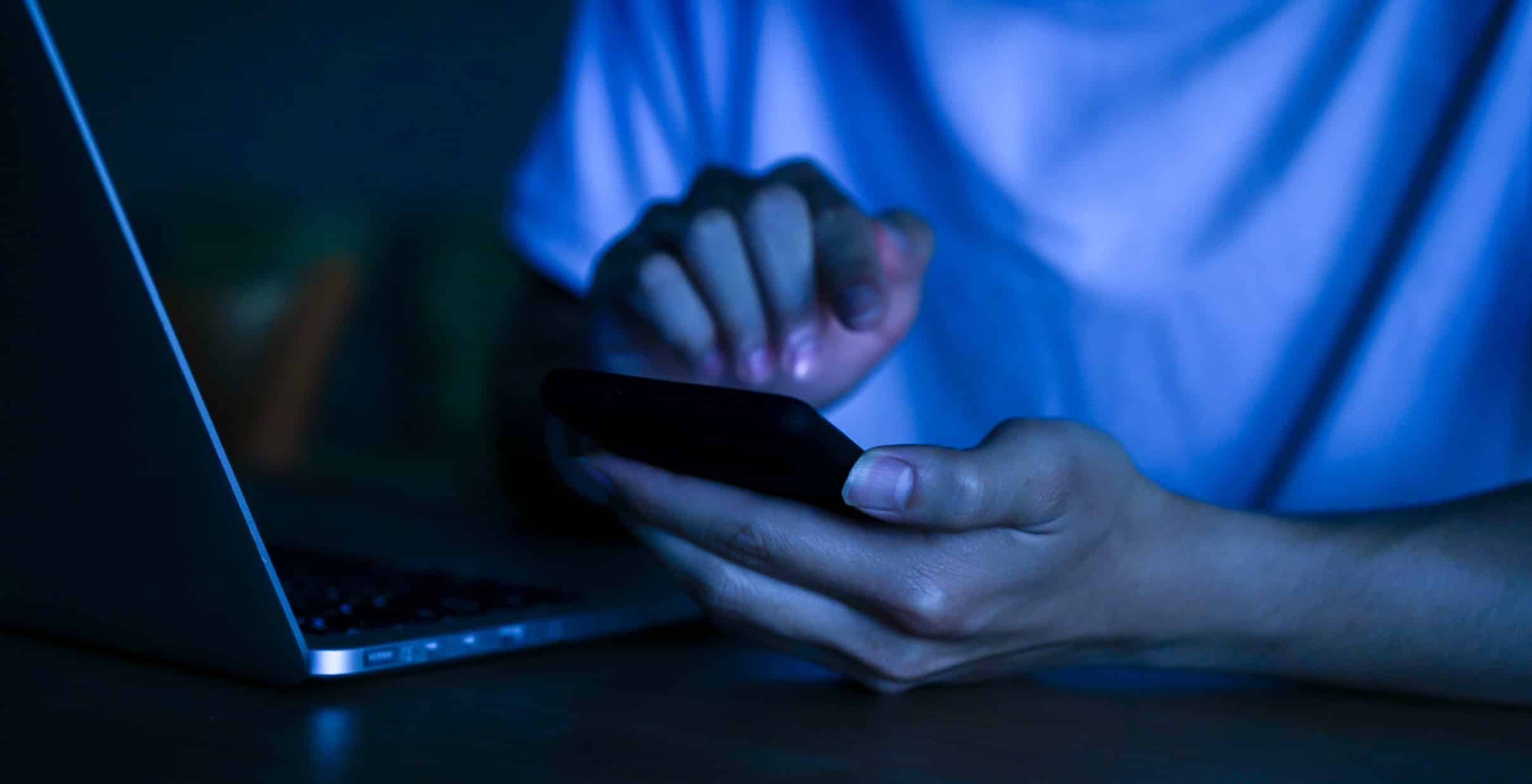 En person använder en smartphone med ena handen och skriver på en bärbar dator med den andra handen i ett rum.