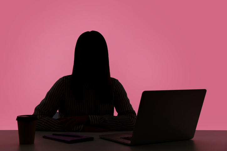 En siluett av en person som sitter vid ett skrivbord med en laptop, en kaffekopp och en surfplatta mot rosa bakgrund.