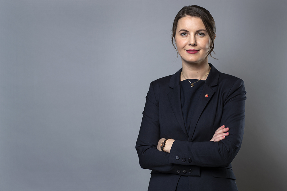 Civilminister Ida Karkiainen i mörk kostym står med armarna i kors mot en grå bakgrund och tittar in i kameran med neutral min.