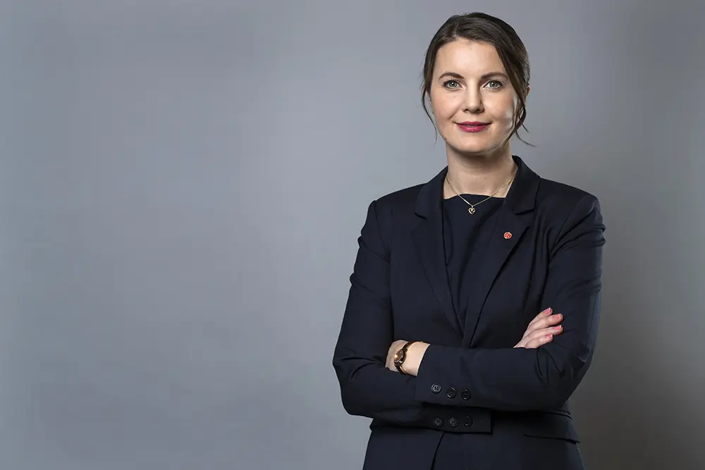 Ida Karkiainen. i mörk kostym står med korslagda armar mot en grå bakgrund.
