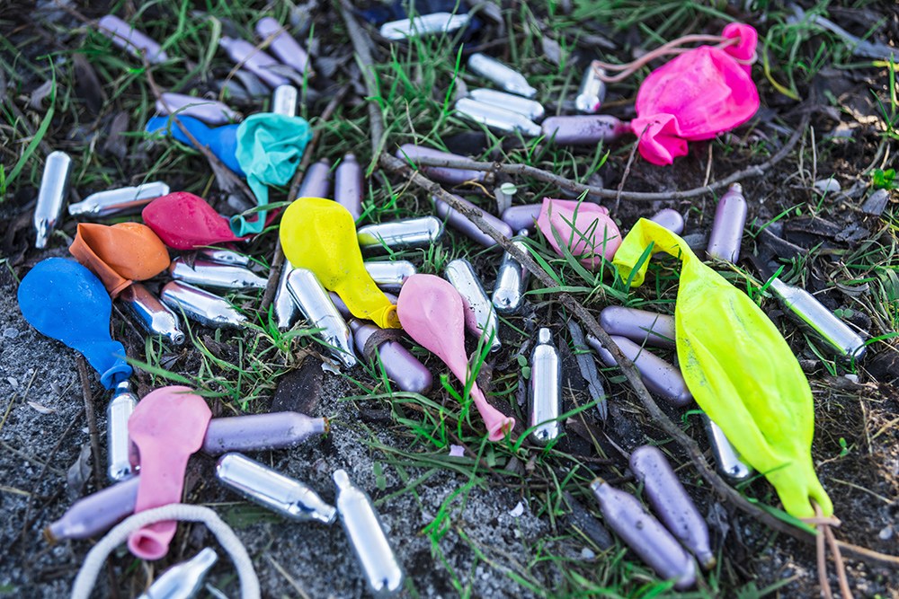 Uppblåsta ballonger och tomma patroner för lustgas ligger utspridda på gräs och jord.
