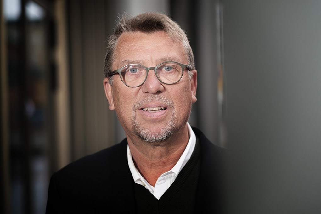 Tapio Salonen med kort ljust hår, glasögon och skägg, i svart jacka och vit skjorta, tittar mot kameran.