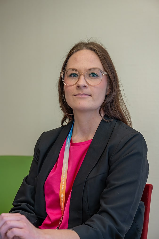 Emelie Norgren med glasögon, svart kavaj och rosa topp sitter inomhus och tittar in i kameran.