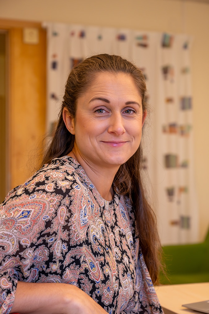 Frida Johansson med långt brunt hår och mönstrad blus tittar i kameran och ler.