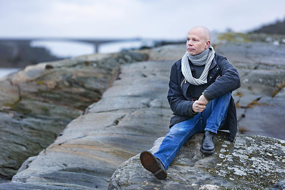 Lasse Mattila sitter på en klippa i ett skärgårdslandskap, med en bro synlig långt bort i bakgrunden