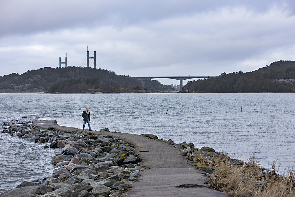 Lasse Mattila långt bak i bild, vandrades på en smal asfaltsväg vid en kust, långt i bakgrunden en bro.