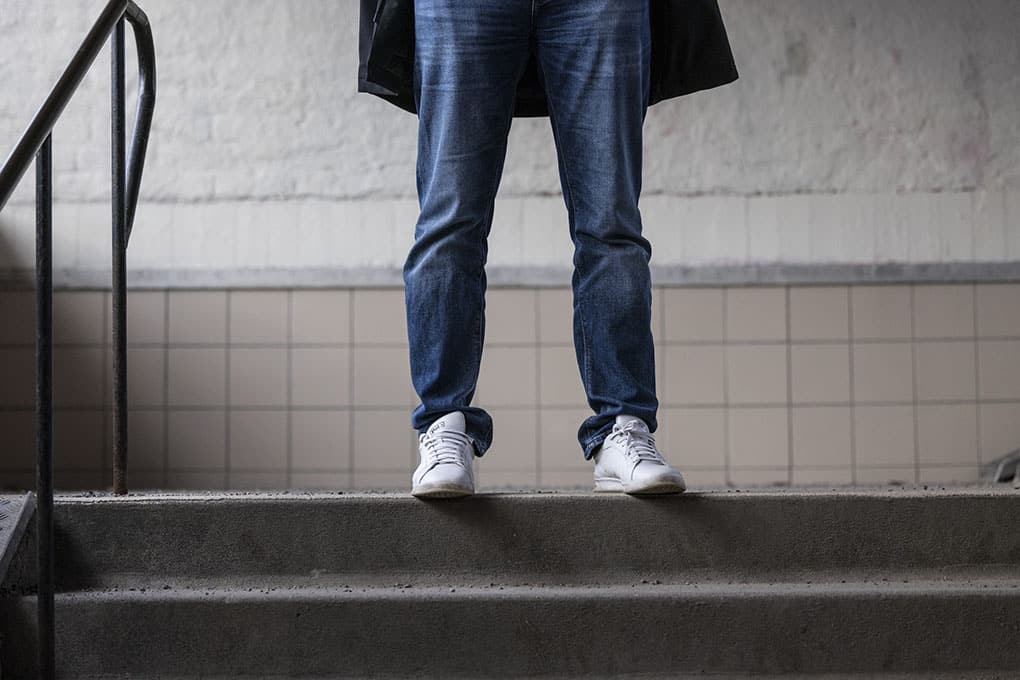 Bild på en man i halvfigur där bara benen syns, klädd i jeans och vita sneakers. Står i vad som ser ut att vara en trappuppgång.