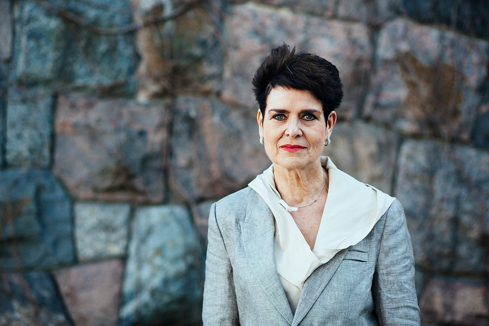 Porträttbild på Ullakarin Nyberg fotograferad framför en stenvägg i svart kort hål och ljusgråkavaj och rött läppstift.