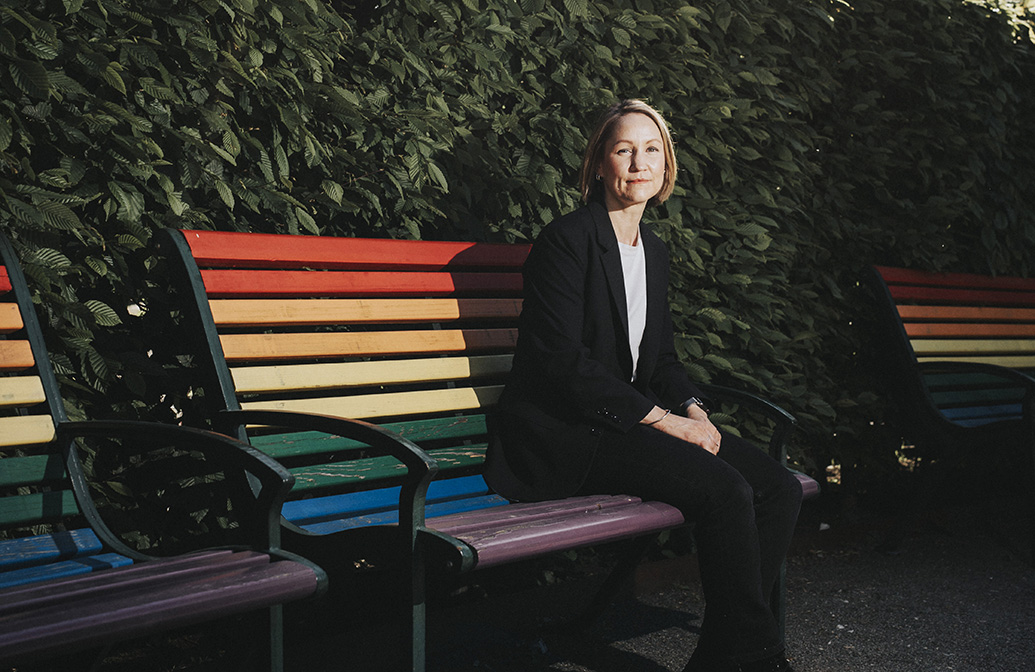 Erika Sundström sitter på en parkbänk i regnbågens alla färger framför en stor grön buske.