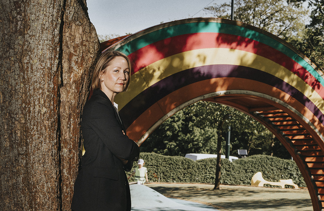 Erika Sundström lutar sig mot ett träd framför ett konstverk föreställande regnbåge.