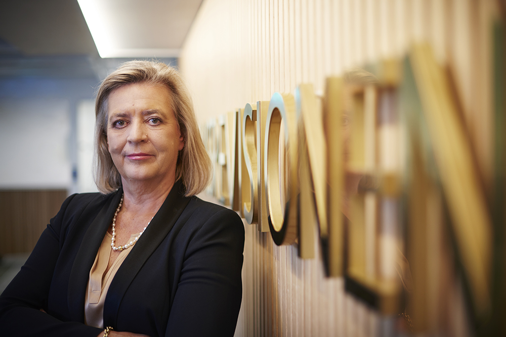 Riksrevisor Helena Lindberg står framför Riksrevisionens skylt inne i myndighetens lokaler.