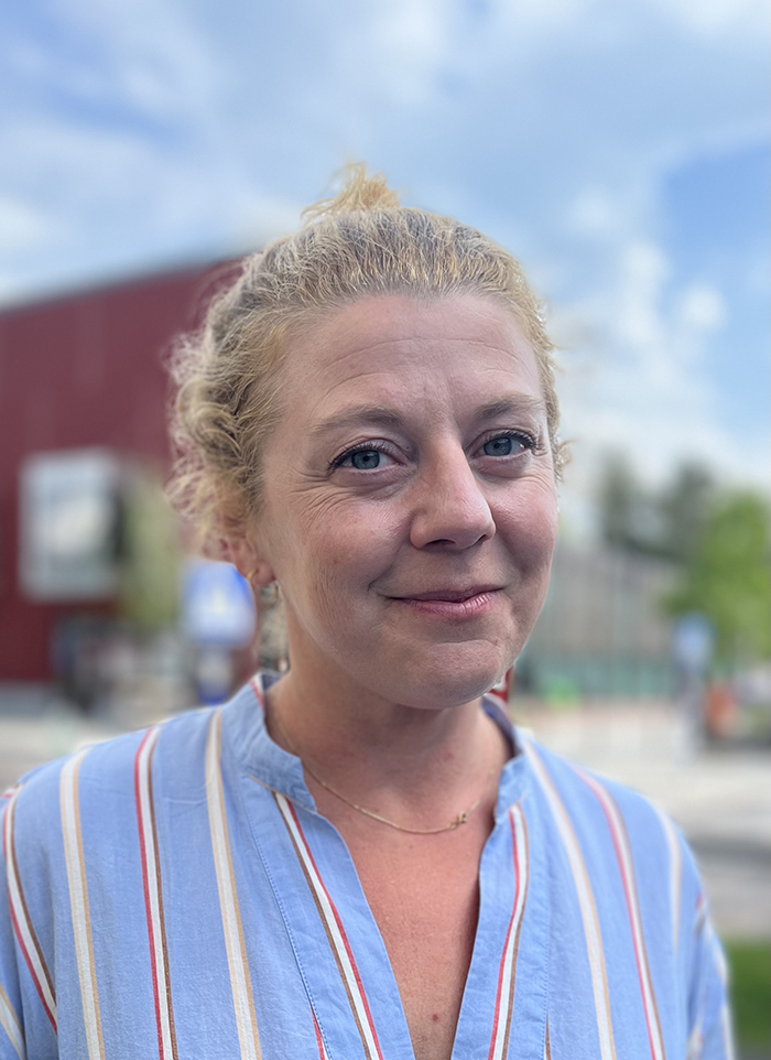 Johanna Morgensterns med lockigt blont hår i en slarvig knut, iklädd blåvit randig skjorta, står utomhus framför en suddig byggnad och himmel.