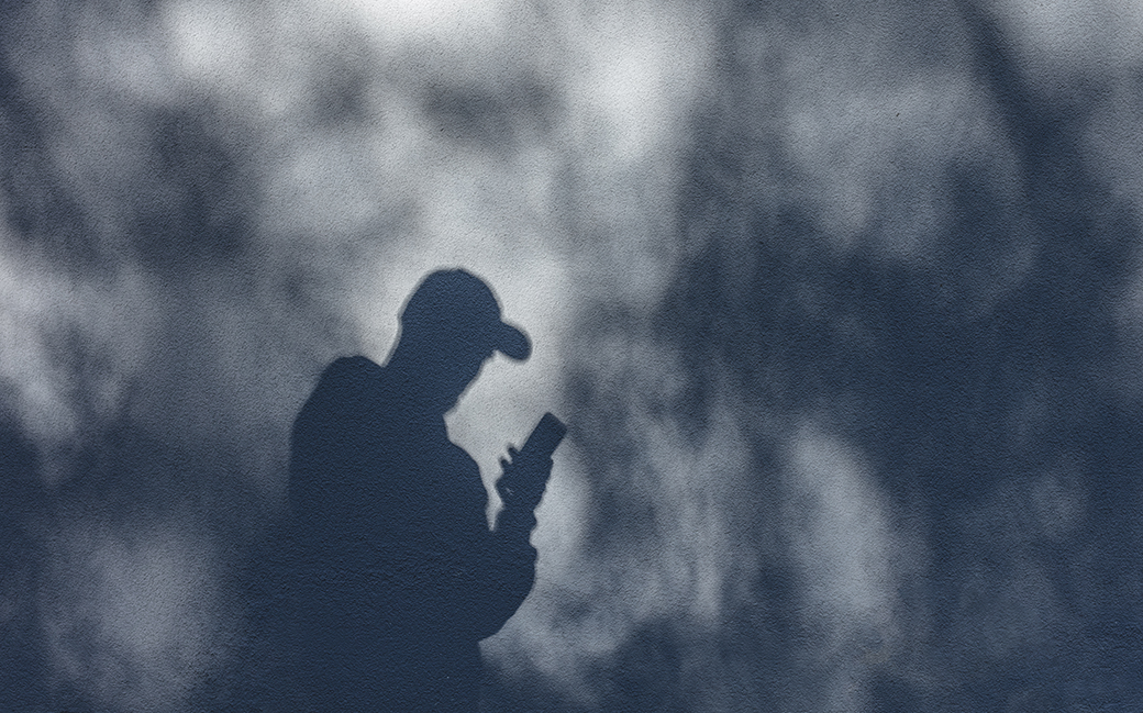 En svart siluett av en person i keps som står i och tittar i sin telefon i en rökig omgivning.