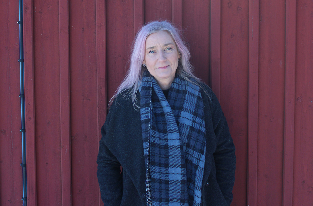 Porträttbild på forskaren Anna-Karin Åkerman i halvprofil, ståendes framför en rödmålad träfasad.