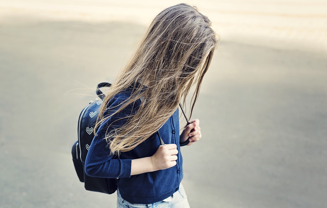 Ett skolbarn med ansiktet sänkt och dolt av sitt långa år promenerar fram med händerna knutna kring ryggsäckens axelremmar.