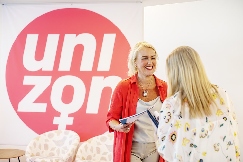 Olga Persson tillsammans med en kollega inne i Unizons lokaler, ståendes framför en stor bild av förbundsloggan.
