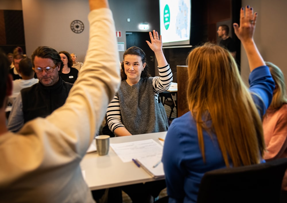 Flertal personer med uppsträckta armar sitter runt bord i en konferenssal.