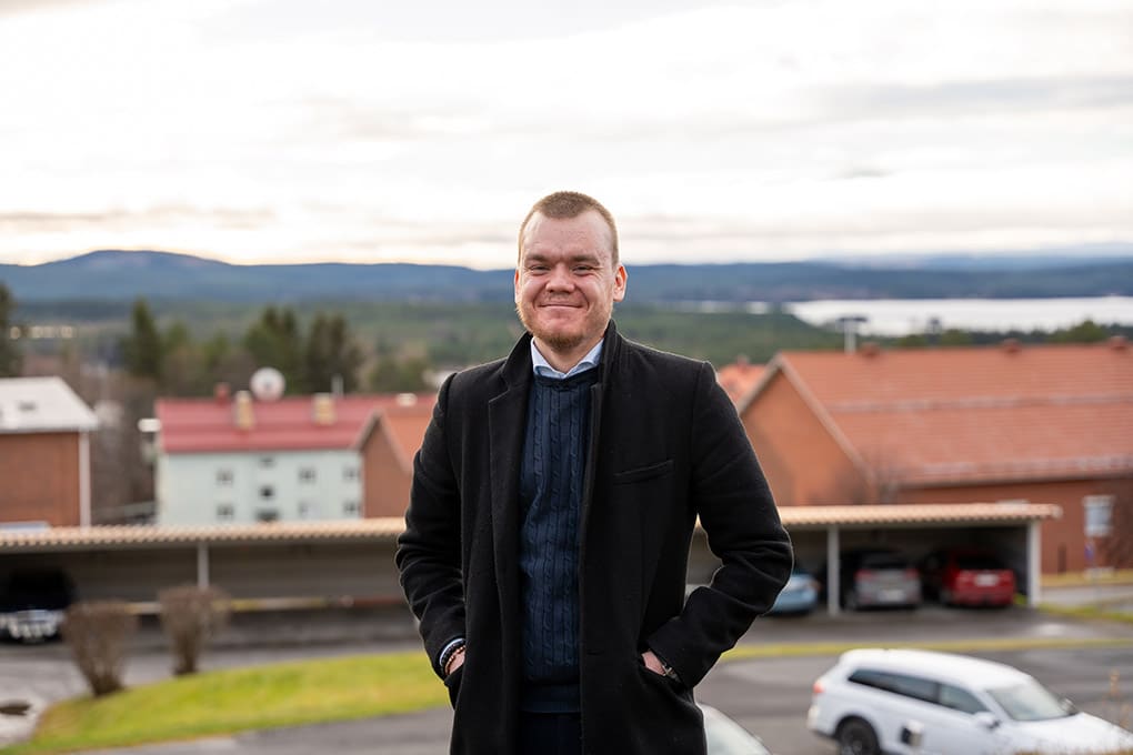 Socionomen Oskar Konradsson står i halvprofil mitt i bild med en vy över Vilhelminas landsbygd i bakgrunden.