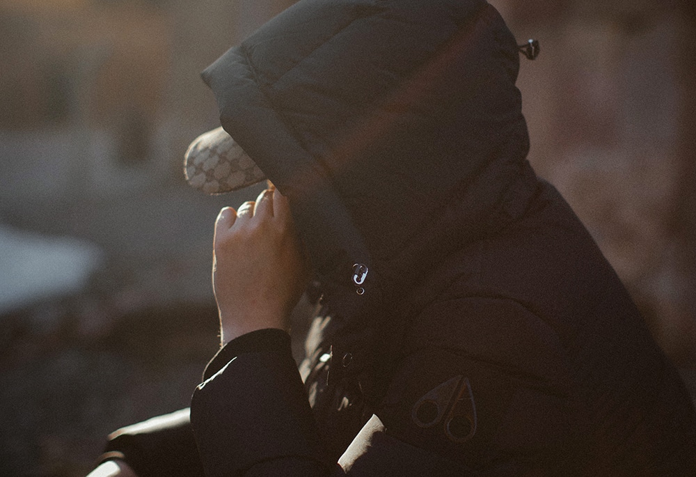 En anonymiserad person sitter och tillsynes pratar i telefon, dold av luvan från en svart dunjacka.