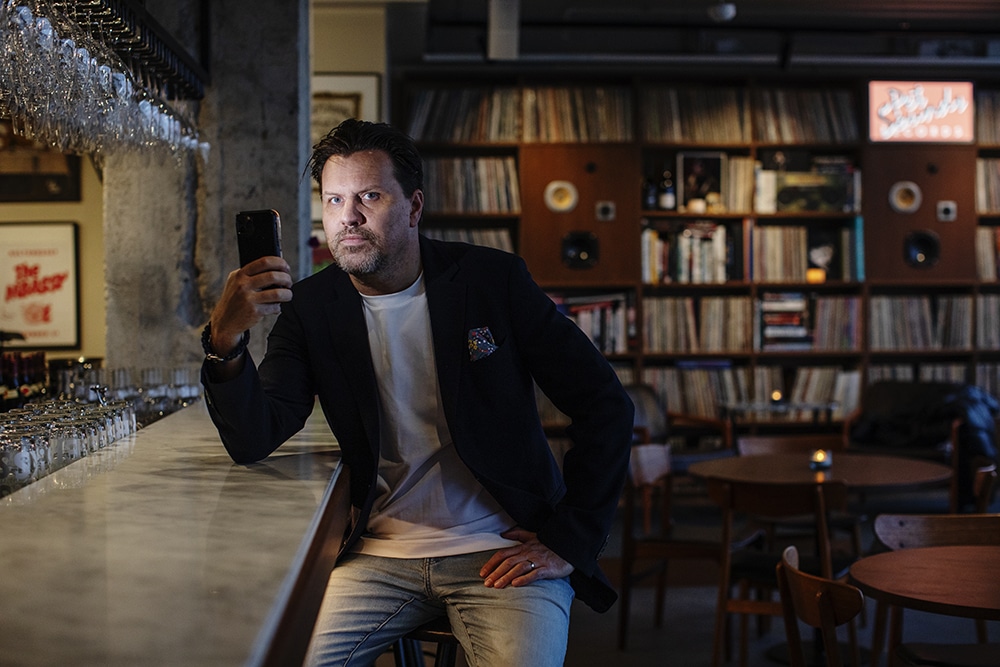 Henrik Pallin sitter på en pall i en bar med sin smartphone riktad mot kameran