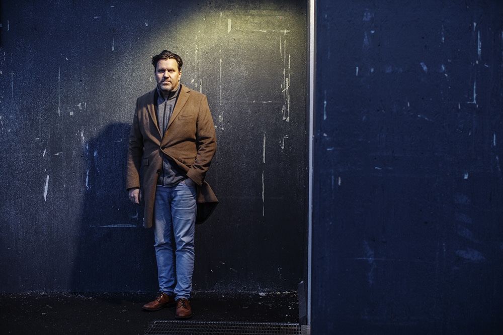 Henrik Pallin står utomhus i en beige överrock, lutad mot en svart vägg, svagt upplyst från sidan av en gatlampa
