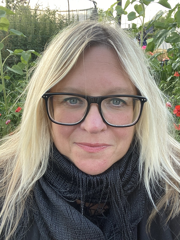 Porträttbild på Sara Kleijsen Åhlander, insvept i en halsduk framför ett buskage.