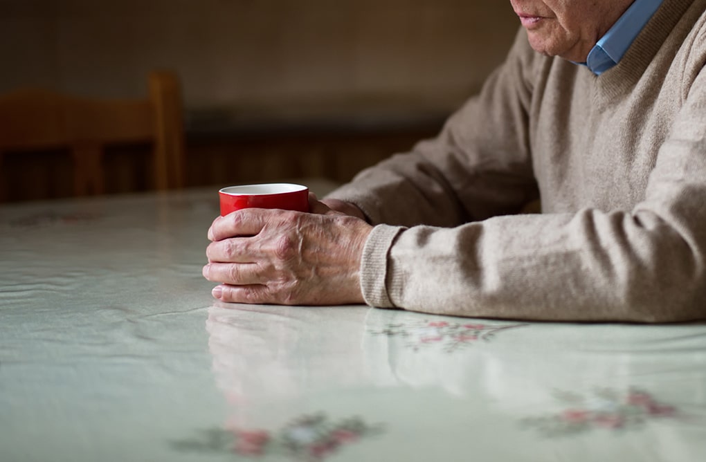 Anonymiserad bild på en äldre person som syns sitta vid ett köksbord med händerna omslutna kring en kaffekopp.