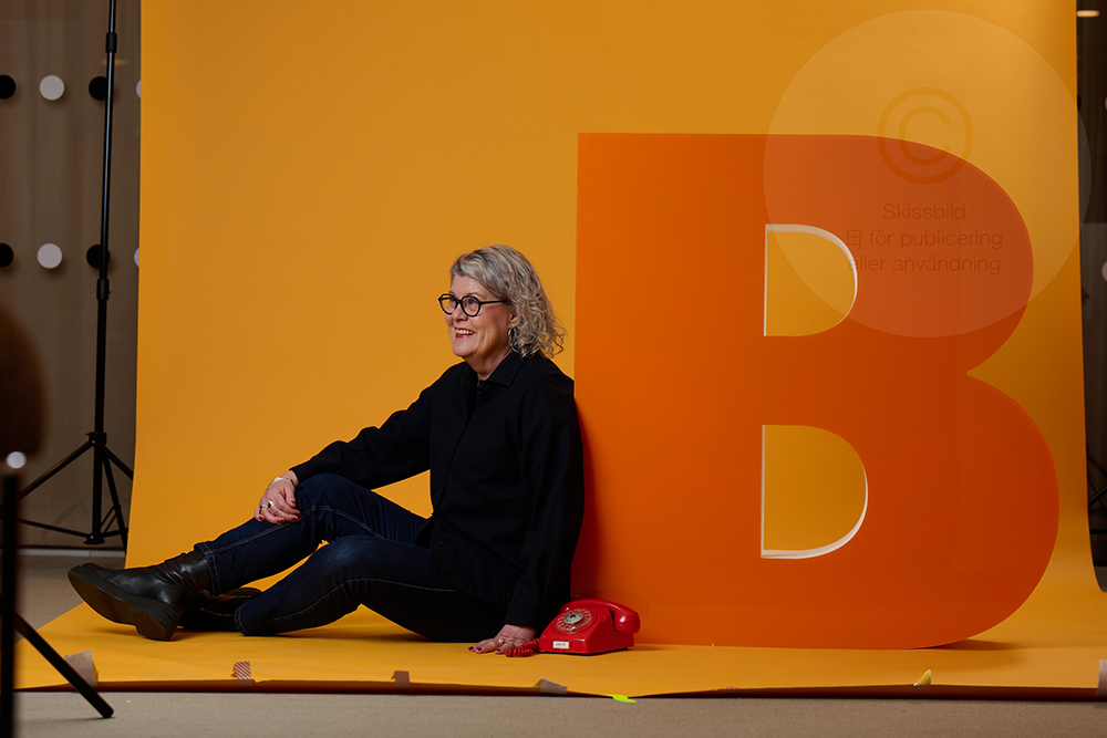 Gunilla Bergsten sitter lutad på ett stort orange B framför en vidsträckt gul fond.